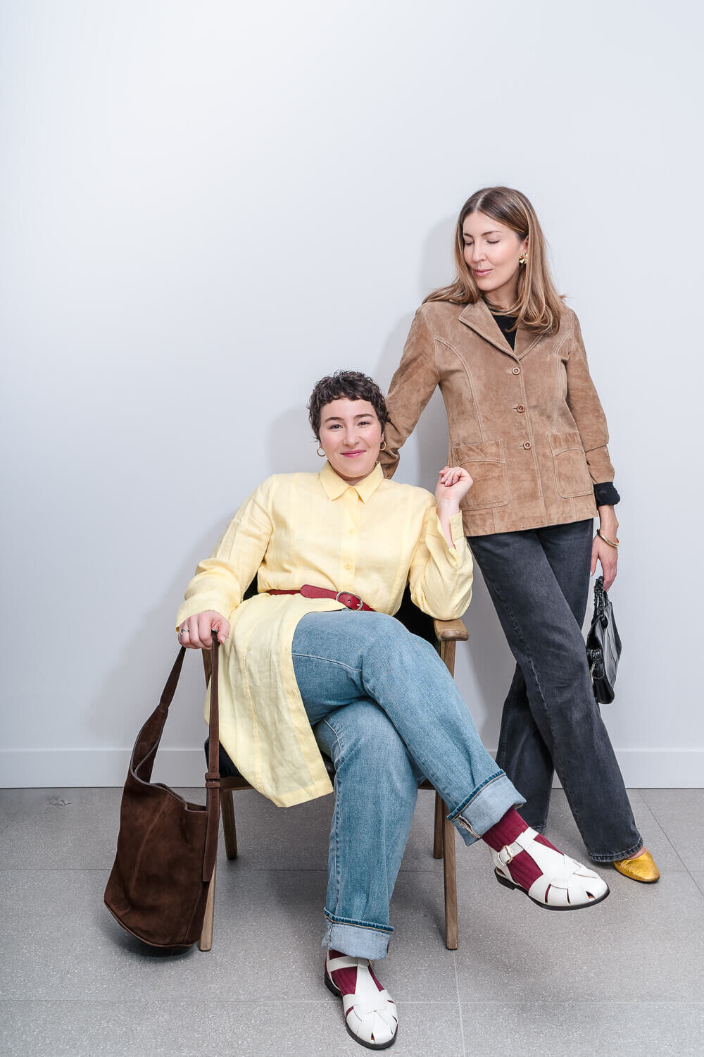 Woman seated in yellow shirt with handbag beside woman standing in tan jacket holding black purse.