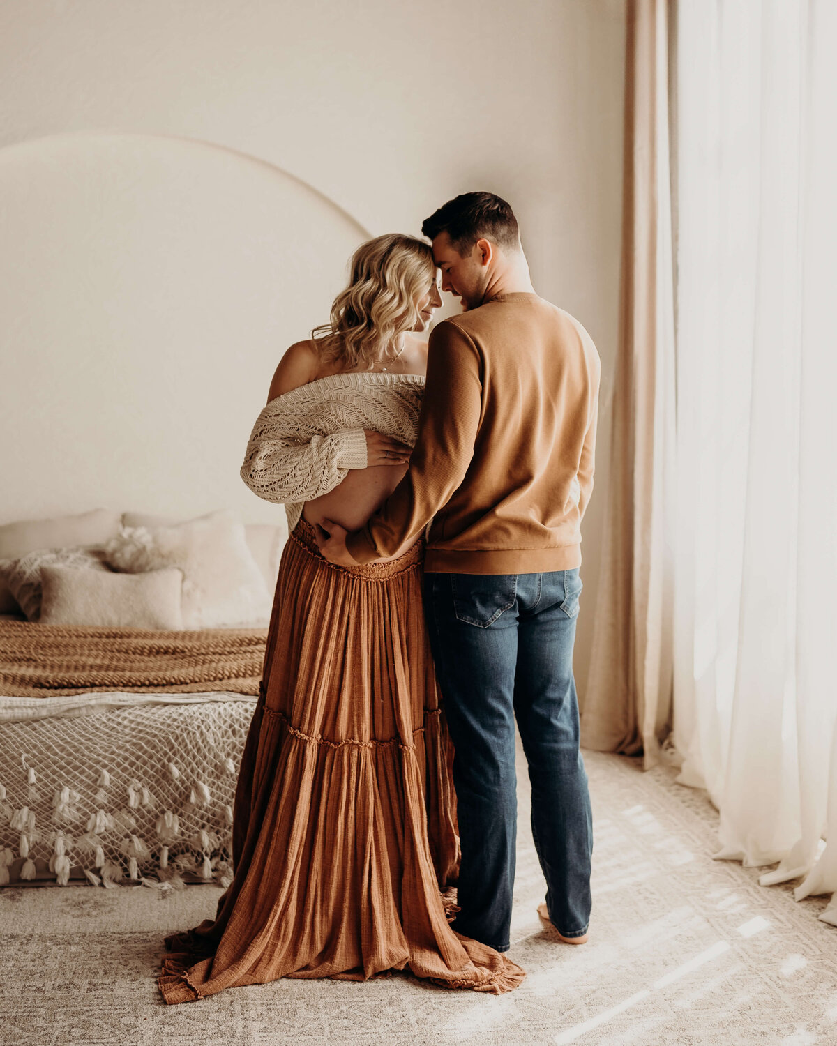 husband and wife stand next to each other foreheads touching while holding her pregnant belly