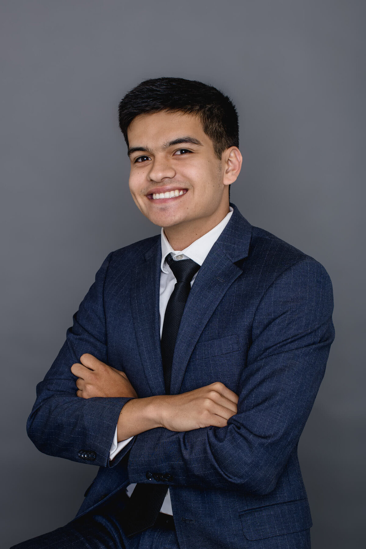 A close-up of a young man with dark hair wearing a navy suit jacket and dark blue tie crossing his arms over his chest in front of a dark grey background.