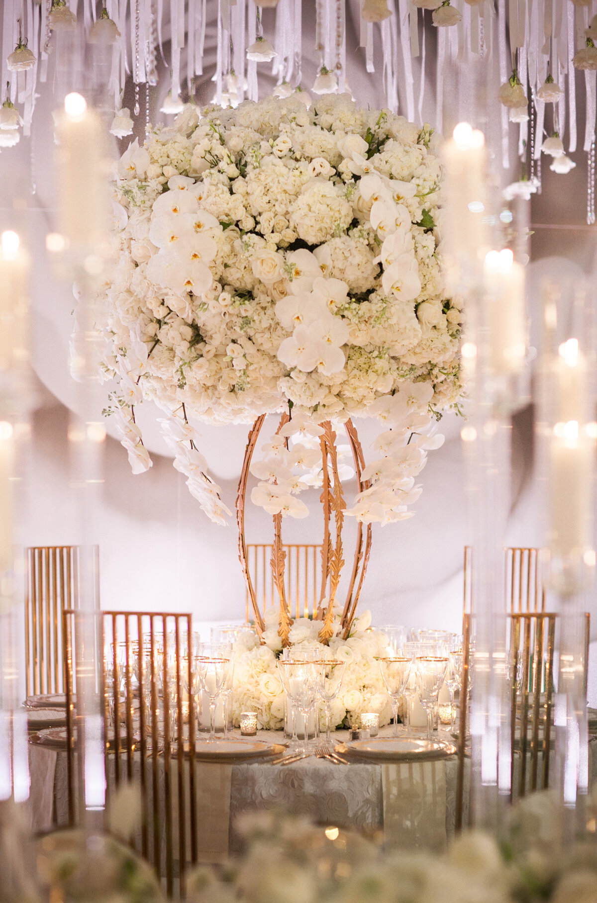 This stunning photo was captured at the iconic New York City location, The New York Public Library, by the talented photographer Christian Oth. The image showcases a lavish wedding table centerpiece adorned with a large array of white flowers, such as roses and orchids, artfully arranged on a gilded structure. Enhancing the display are tall candles, sparkling crystal accents, and sophisticated table settings that harmonize beautifully with the white and gold color palette. Videography for this event was expertly handled by East West Films.