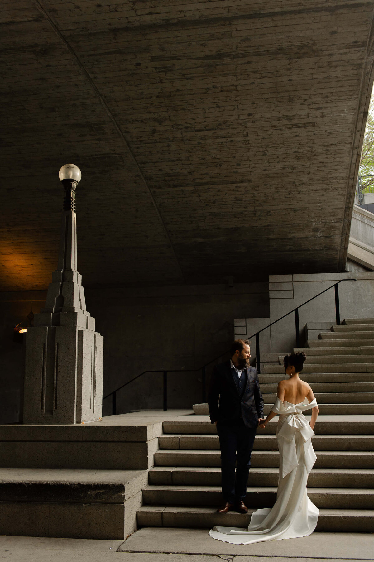 Ottawa wedding photography showing a groom in  blue suit and a bride with a white dress and big bow on the back holding hands on the stairs opposite the Chateau Laurier wedding venue