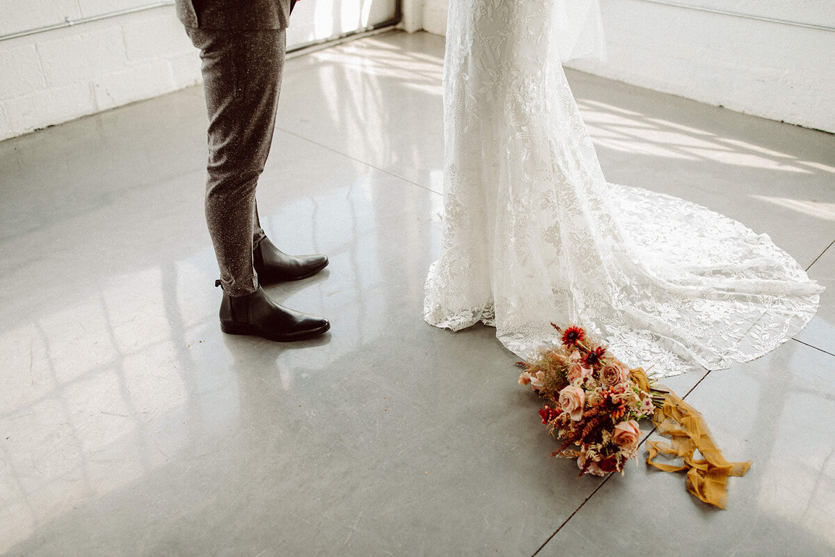 Bride and groom, wearing a gray suit and white wedding gown, stand next to a bouquet on ivory tile.