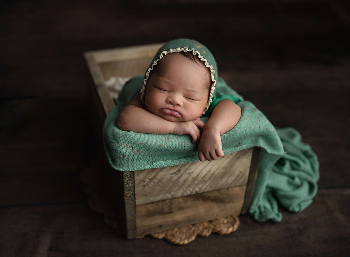 A newborn baby sleeps in a wooden box in a green bonnet taken by a New Orleans Newborn Photographer