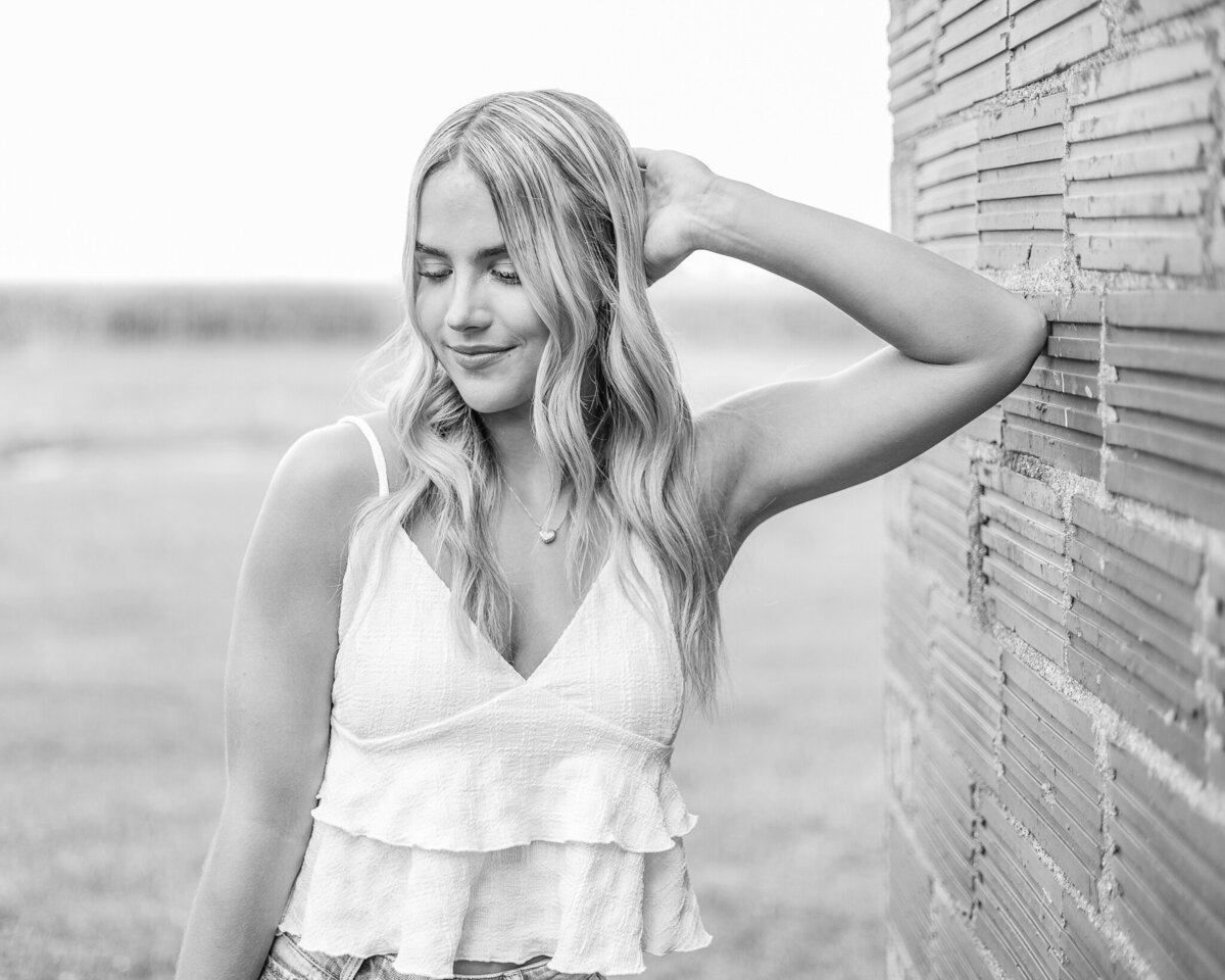 black and white photo of a high school senior female with long hair, looking dow with a soft smile, has one elbow leaned against a brick wall, wearing a spaghetti strap top