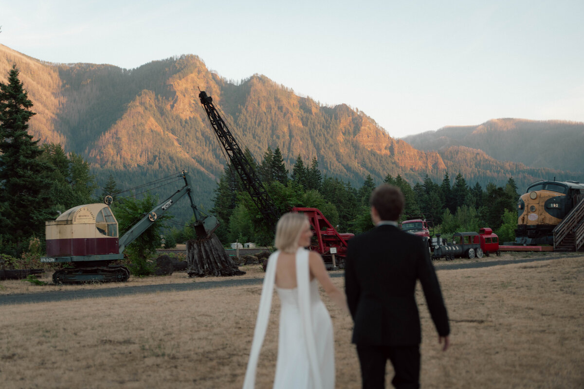 Bride and groom walking toward mountain landscape at sunset with red logging trucks in background