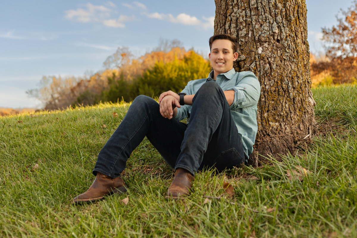 A senior boy sitting against a large tree, smiling while wearing a casual sweater and jeans.