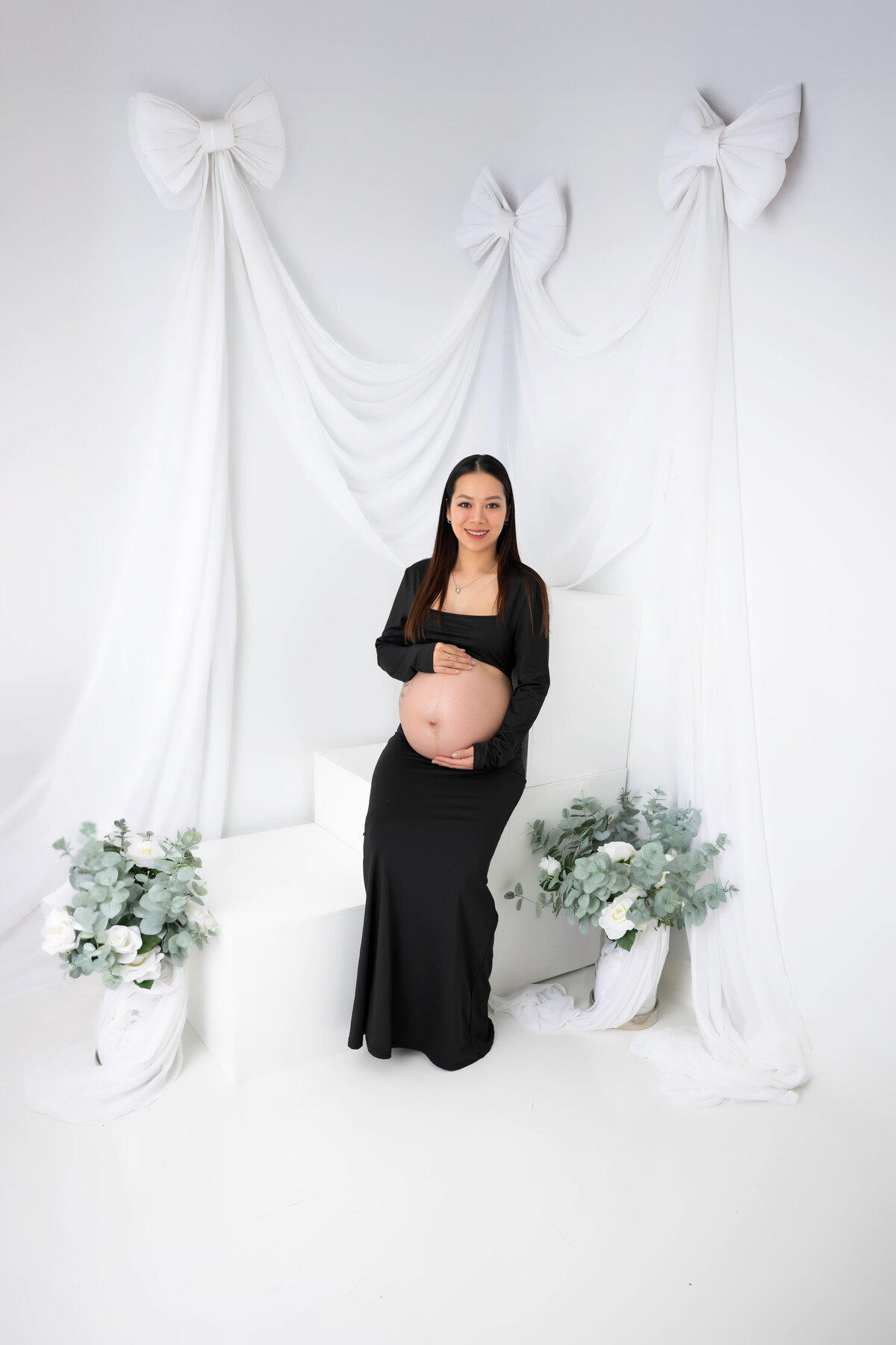 Woman pregnant in black attire sitting on white stairs, embracing her bump.