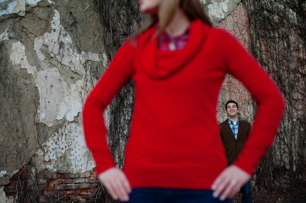 a woman wearing a bright red sweater has her hands on her hips.  Through her arms you can see her fiance in the background