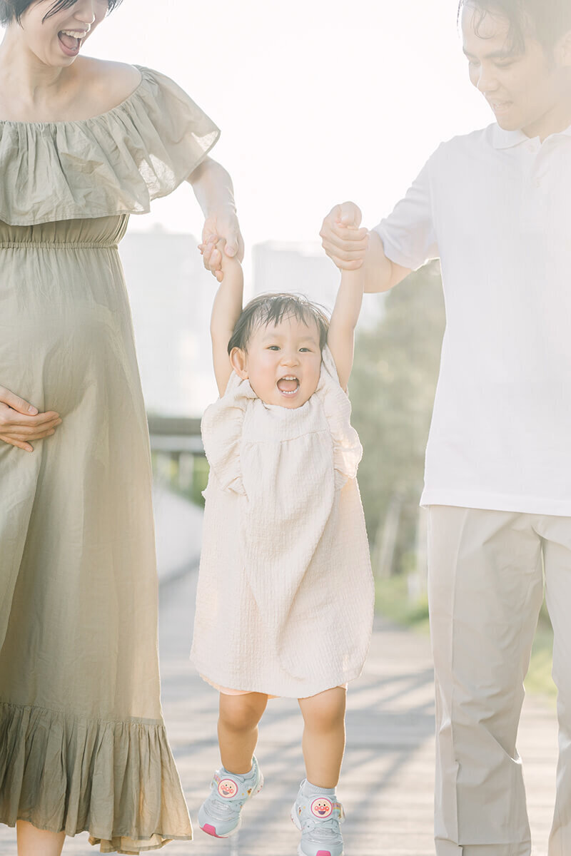 A little girl smiling as her mom and dad take her hand and hold her up in the air. Beautiful family maternity session in Tokyo by Yukika Ishikawa Photography