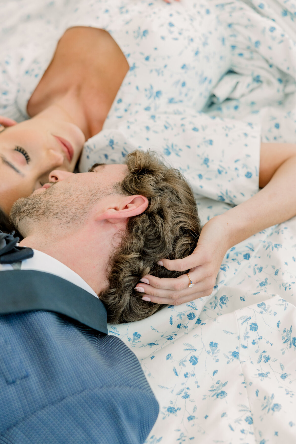 Ivy Rose Barn | A closeup of a bride in a vintage-inspired puff sleeve floral gown and groom in navy tuxedo laying on the train of the bride’s dress. As they cup each others’ faces, you can see the bride’s ring sparkle as she touches the groom’s curly hair.
