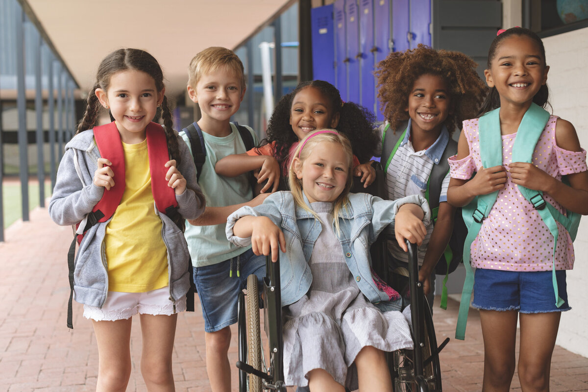 A multi-cultural group of children including a young girl in a wheelchair smiling while in a school hallway