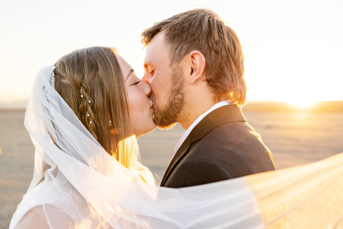 Bride and groom share a kiss as the bride’s veil flows in the wind, with the sunrise in the background.