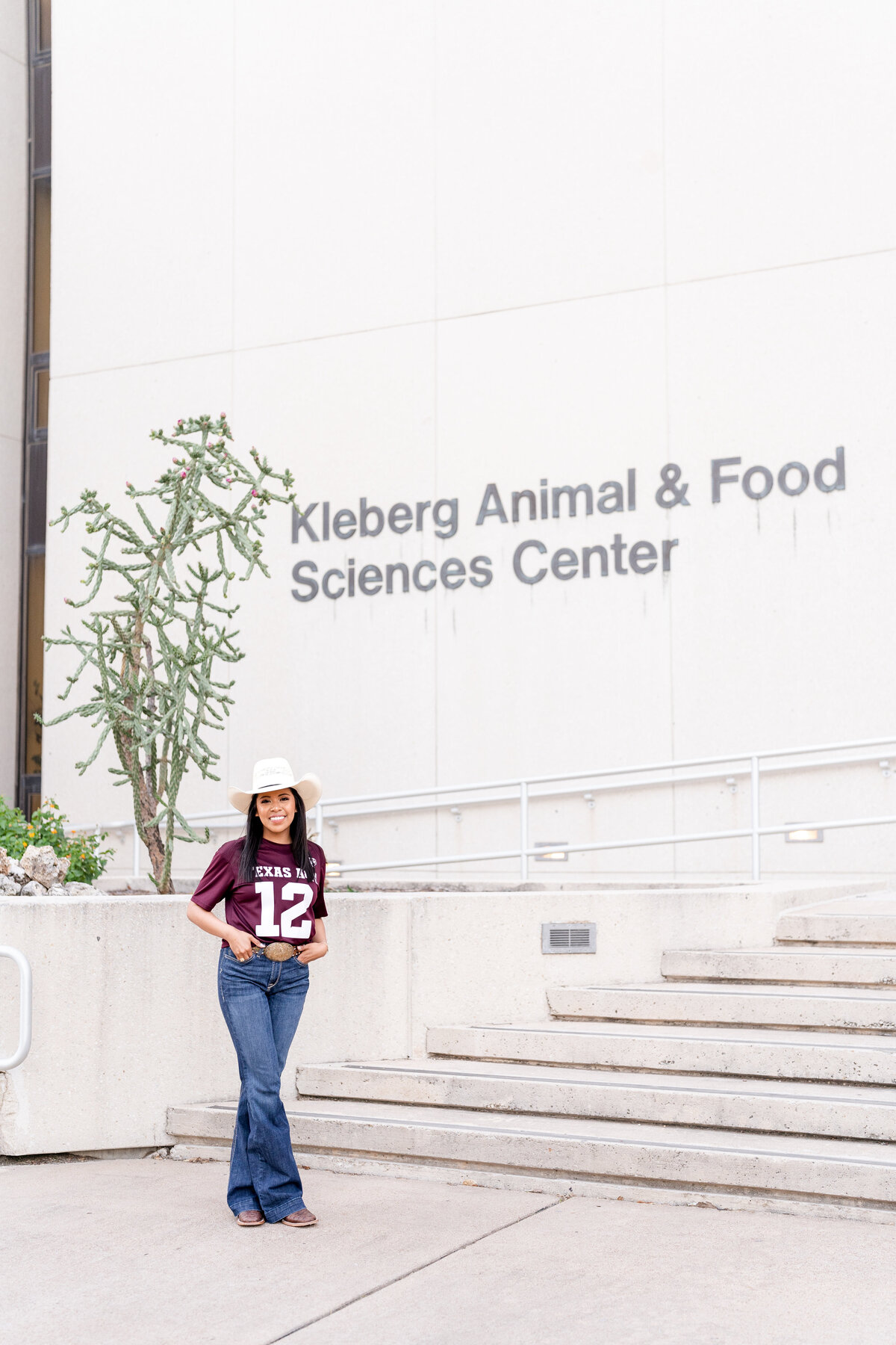 Texas A&M senior girl standing with hands in pockets in front of Kleberg while wearing jeans, cowboy hat and maroon Aggie jersey