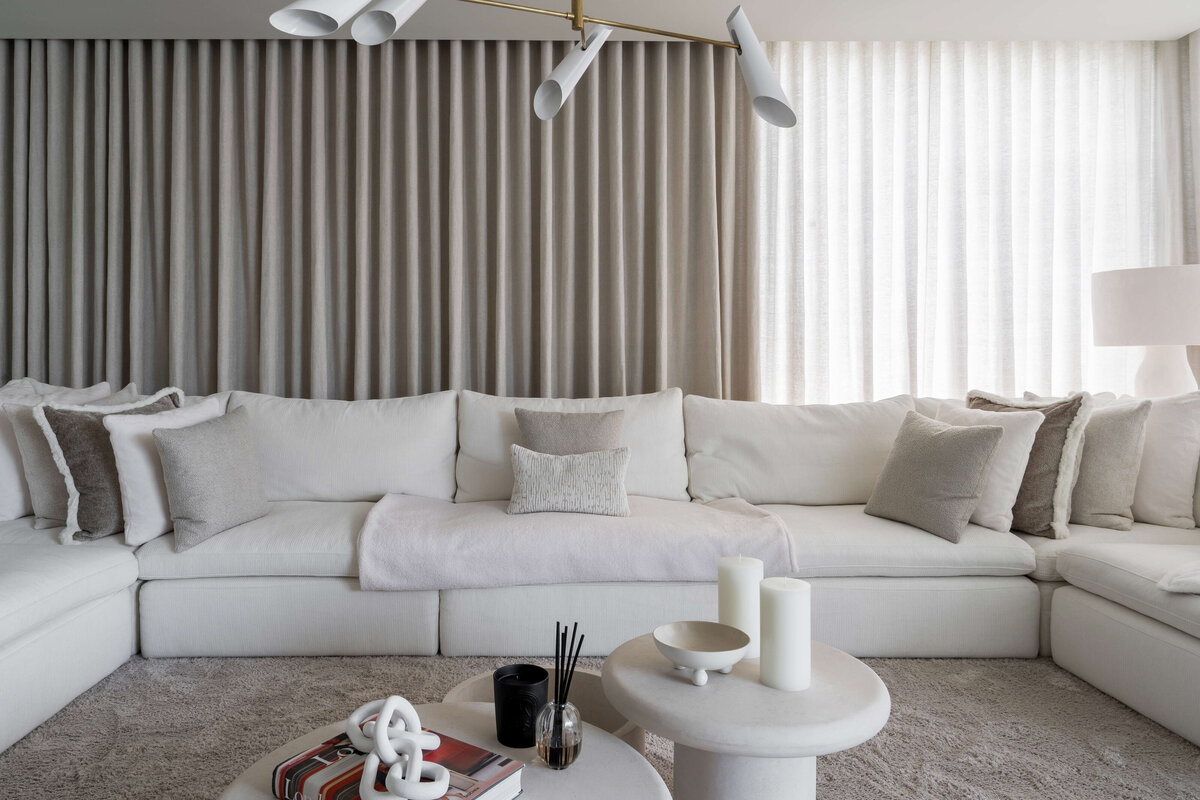 A spacious, modern living area. A large white couch with neutral throw pillows sits on a soft, textured cream rug. Behind the couch are floor-to-celiing cream drapes extending the length of the wall.