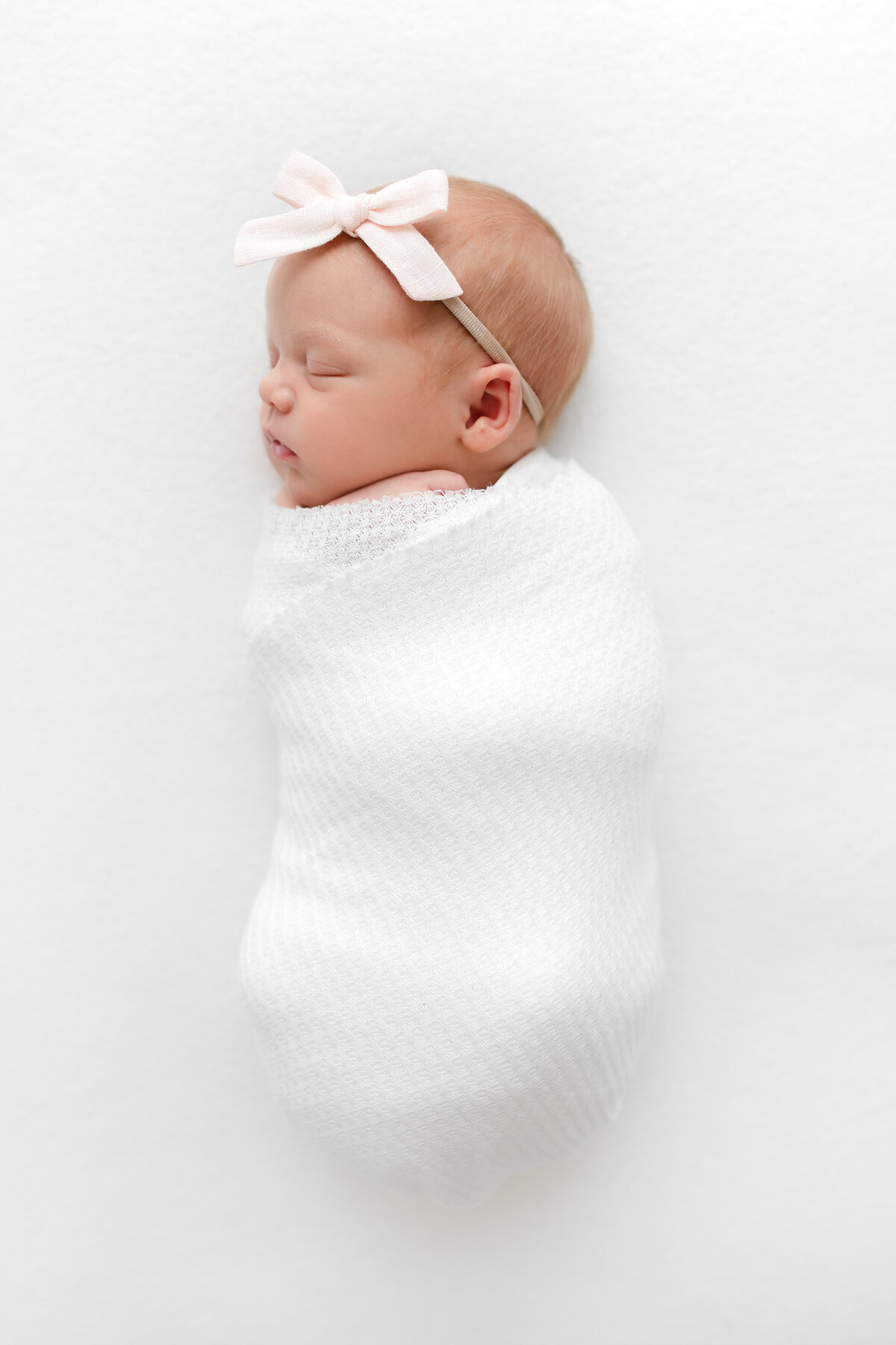 A DC Newborn Photography photo of a baby girl swaddled with a pink bow sleeping on a white blanket