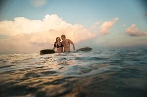 Romantic couple embracing in ocean water at sunset during St Augustine beach wedding photoshoot with surfboard and dramatic clouds