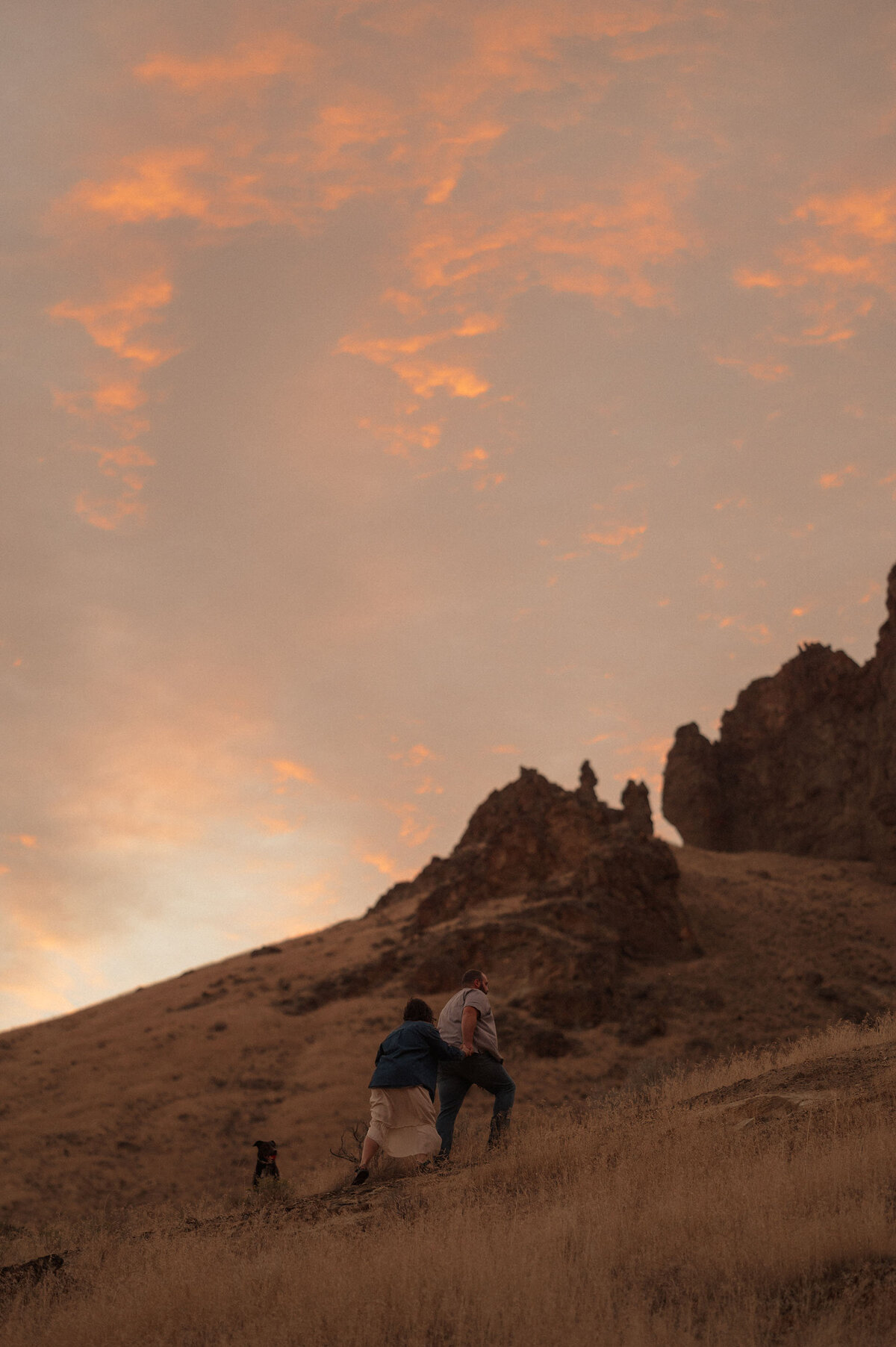 couple walks up a hill with the sunset and jagged rocks in the background under a pink sky in Moab, Utah