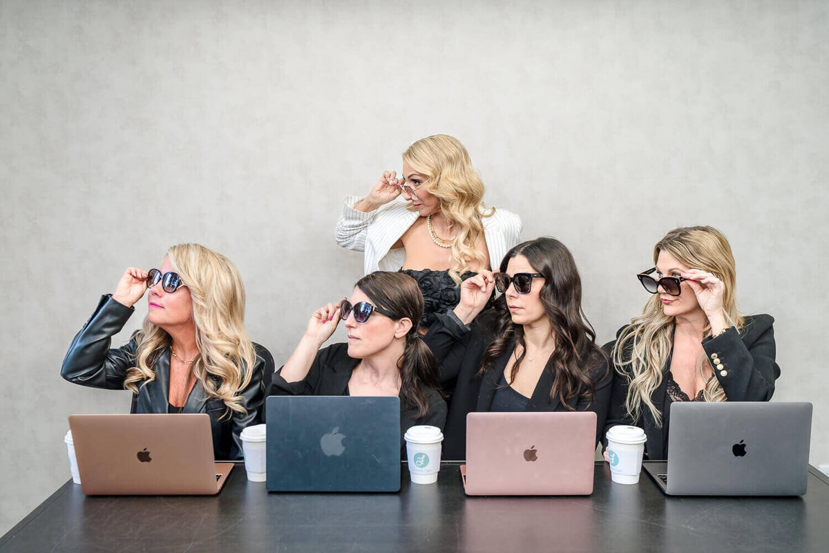 Team of female entrepreneurs in sunglasses looking left with laptops open in front of them.