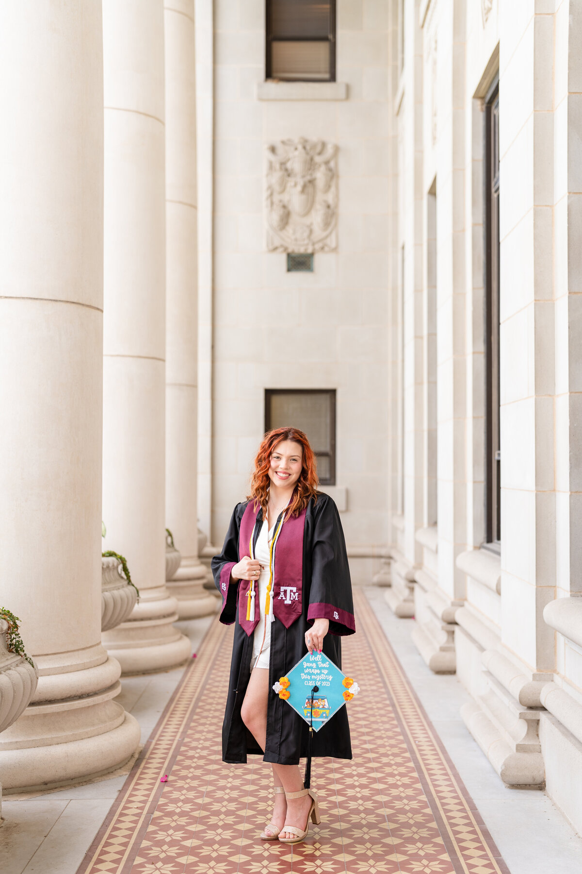 Texas A&M senior girl wearing gown and stole and holding decorated cap and smiling in the columns of the Administration Building