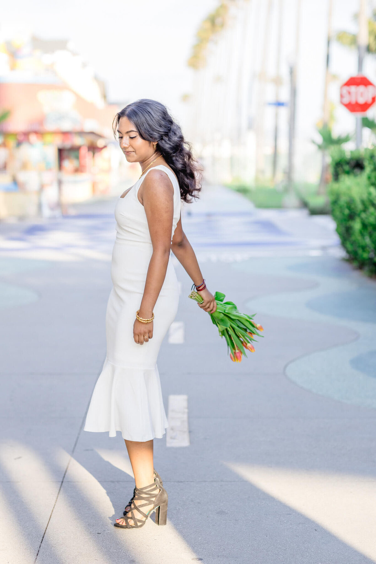 A woman in a white dress stands on a patterned walkway, holding a bouquet of tulips behind her back. She has long, wavy hair and wears high-heeled sandals. The background features blurred buildings and palm trees.