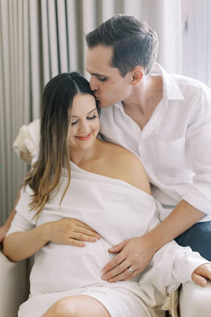 Mommy touching baby bump while daddy kisses her head lovingly.