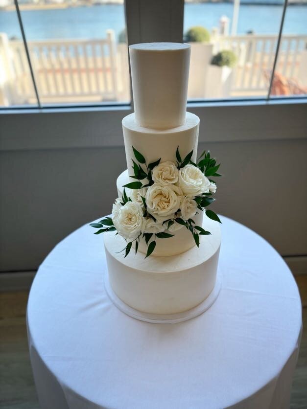 A four tier wedding cake sits on a low cocktail table with a white tablecloth. The cake is all white smooth frosting, and has a cluster of white flowers and greenery in the front middle of the cake.