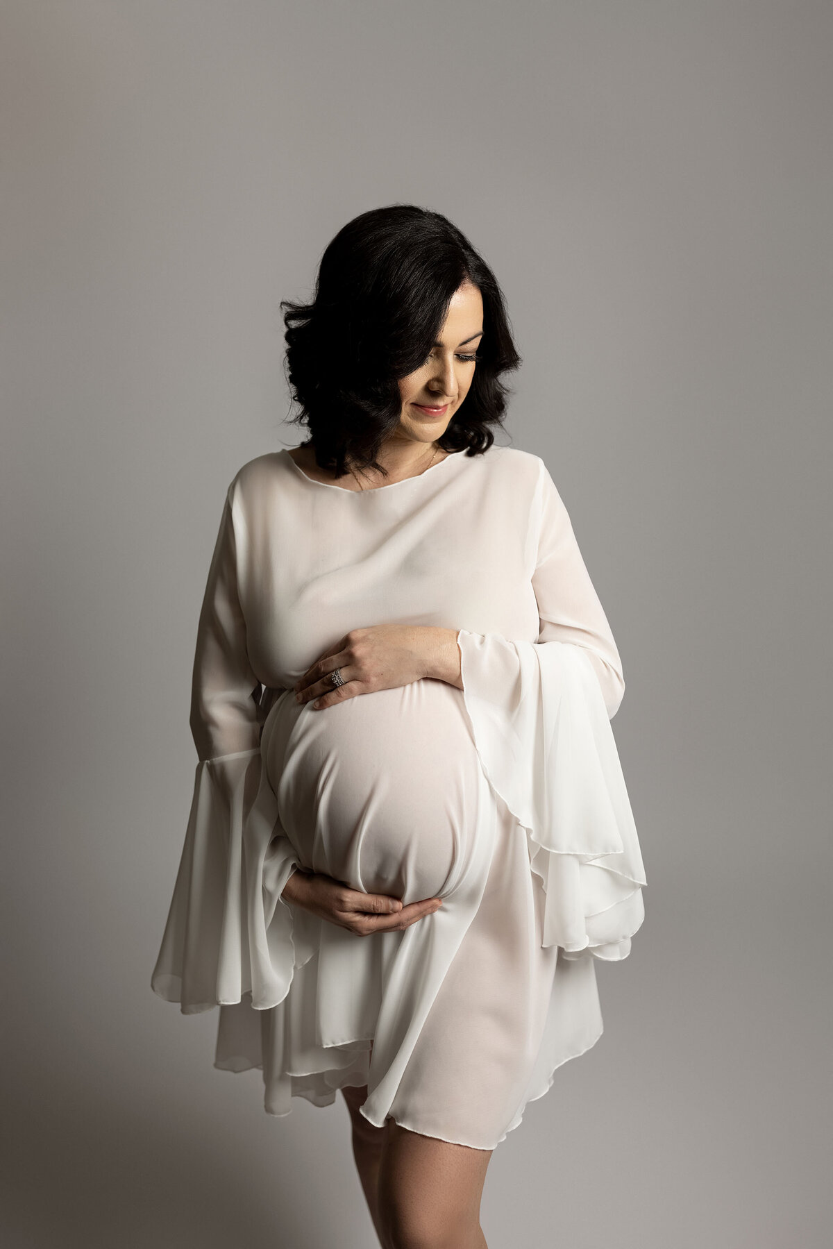 Maternity studio portrait of expectant mom shot by top london, ontario maternity photographer Ogg Photography. Mom is standing in a flowy knee-length white dress with one hand on top and the other above her baby bump. She is looking down and smiling at her belly. Side profile image of mom with shoulder-length wavy black hair.