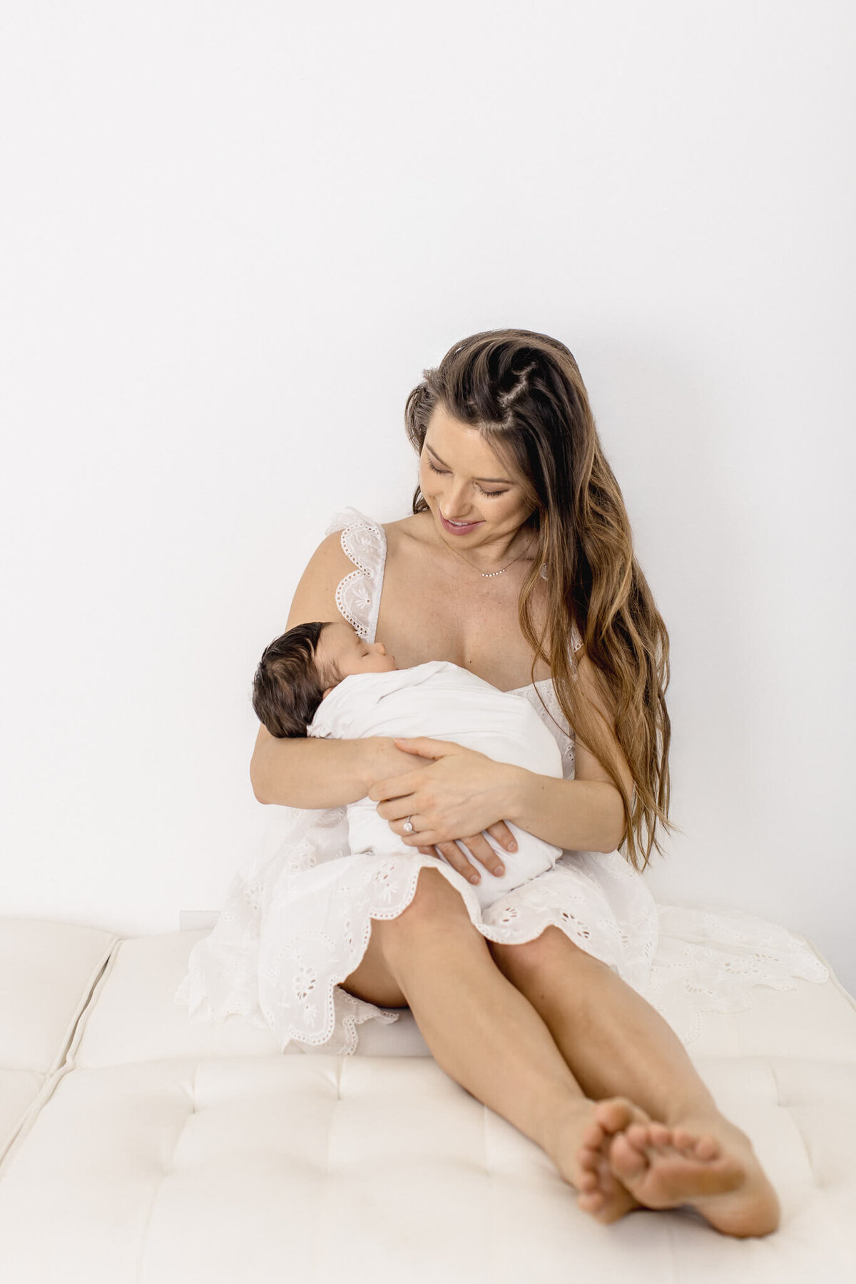 A woman in a white dress sits on a bed, cradling a baby wrapped in a white blanket. She gazes lovingly at the child, with a peaceful expression. The background is a simple white wall.