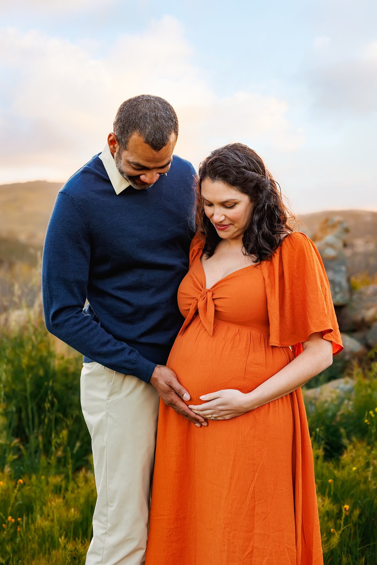 Husband and wife holding pregnant belly and looking down while standing in a lush field