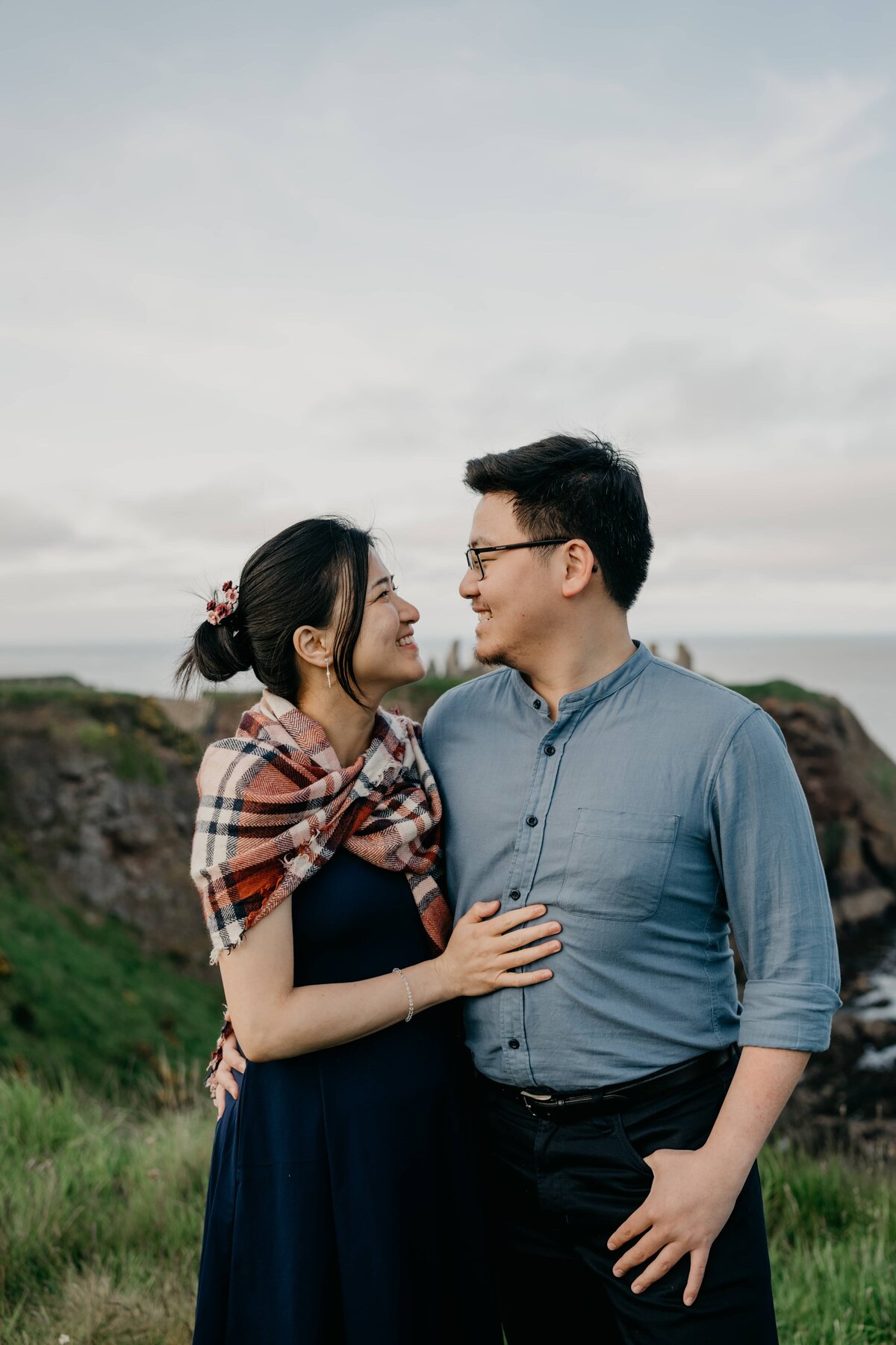 An engaged couple stand with their bodies pressed together and smile lovingly at each other the Aberdeenshire coastline during a photography session.