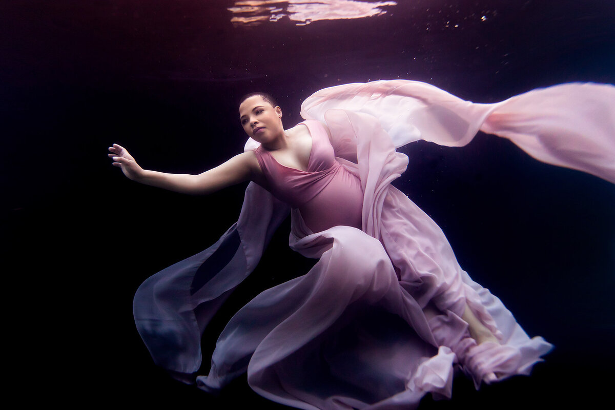 A woman elegantly floating underwater in a flowing pink gown, her arms gracefully extended as the fabric billows around her. The deep black background contrasts beautifully with the delicate pastel tones, creating a mesmerizing sense of calm and poise.