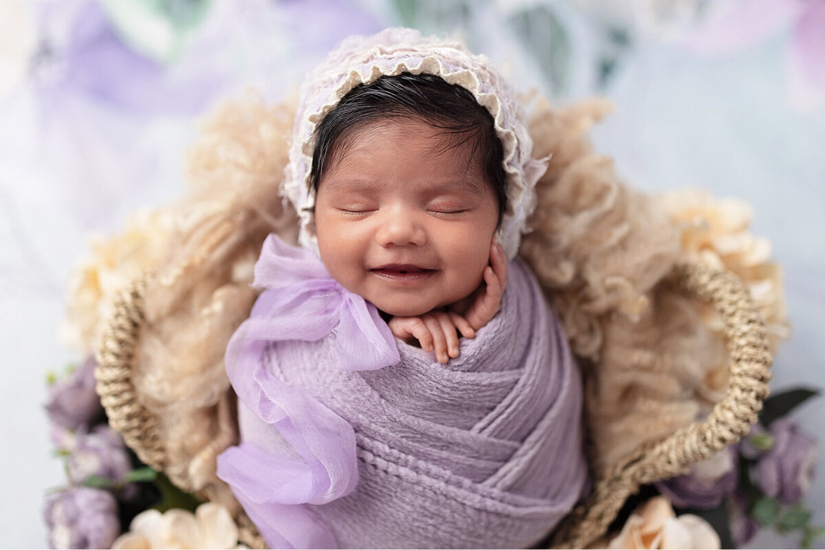 Newborn girl smiling while wrapped in lavender on a soft textured backdrop.