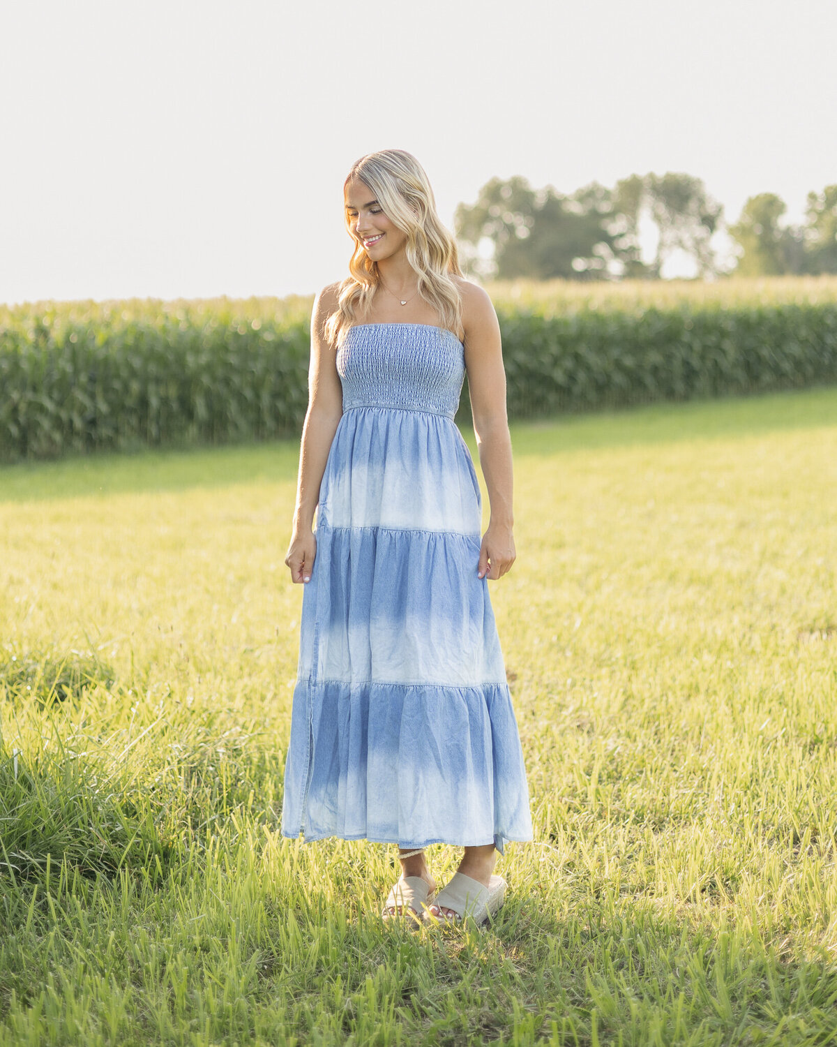 High school senior female with long blonde hair, wearing a blue and white long dress, standing in the grass, with a corn field in the background, pinching the sides of her dress, looking down and smiling