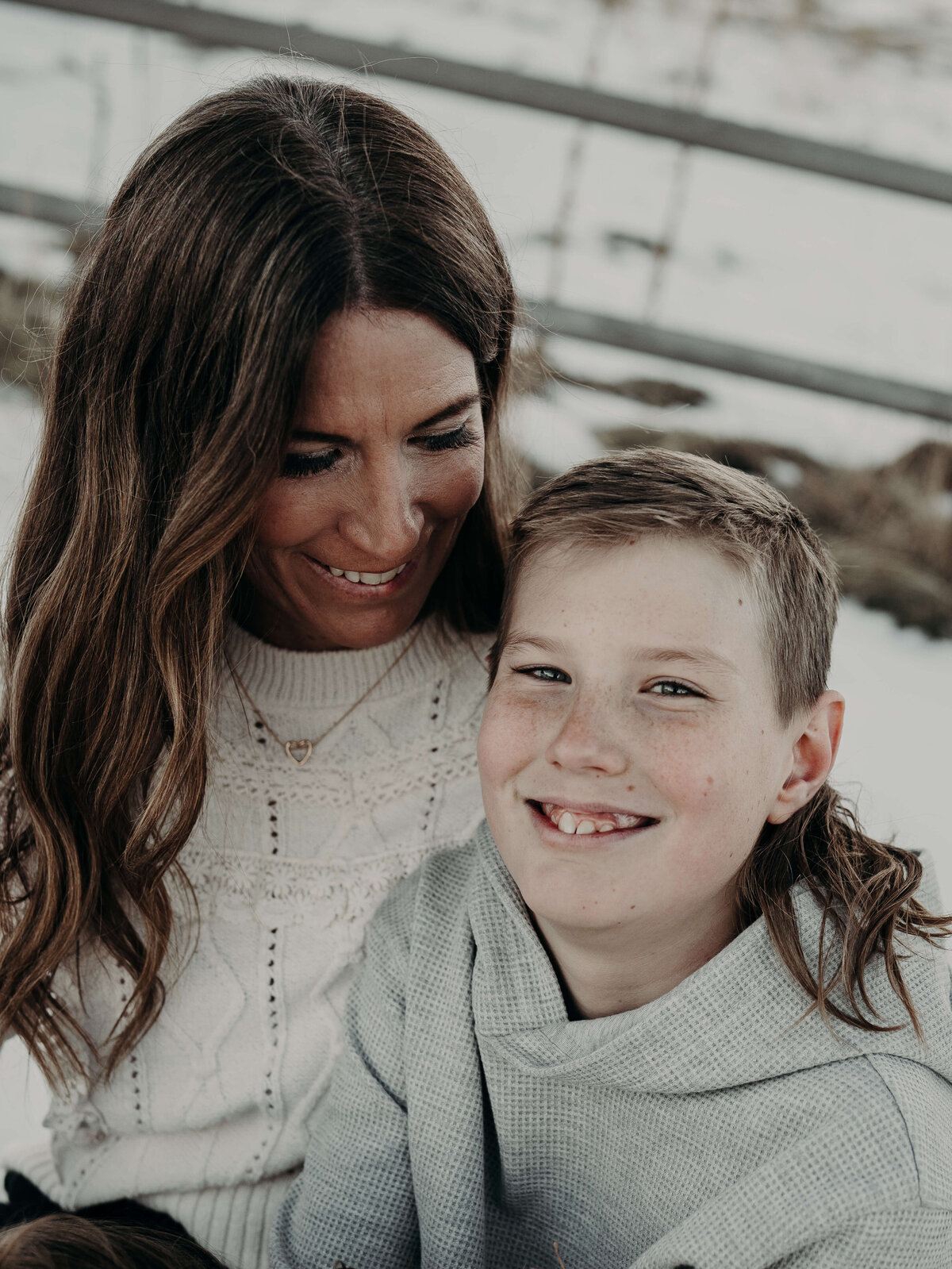 A mother smiles down at her son during a winter photography session, while he smiles directly at the camera.