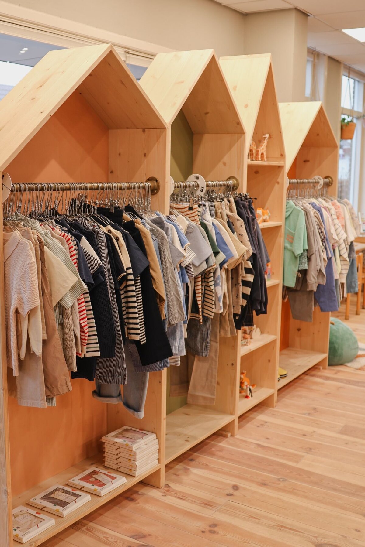 Kinderkleding hangt aan metalen stangen in huisvormige houten rekken in een winkel met een warme, houten uitstraling.
