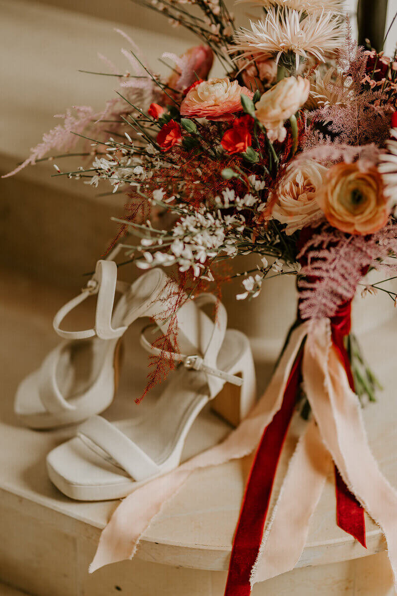 Bouquet de mariée rose et rouge disposé sur des marches en pierres et chaussures à talons blanches. Mise en scène par Laura Termeau photographie.