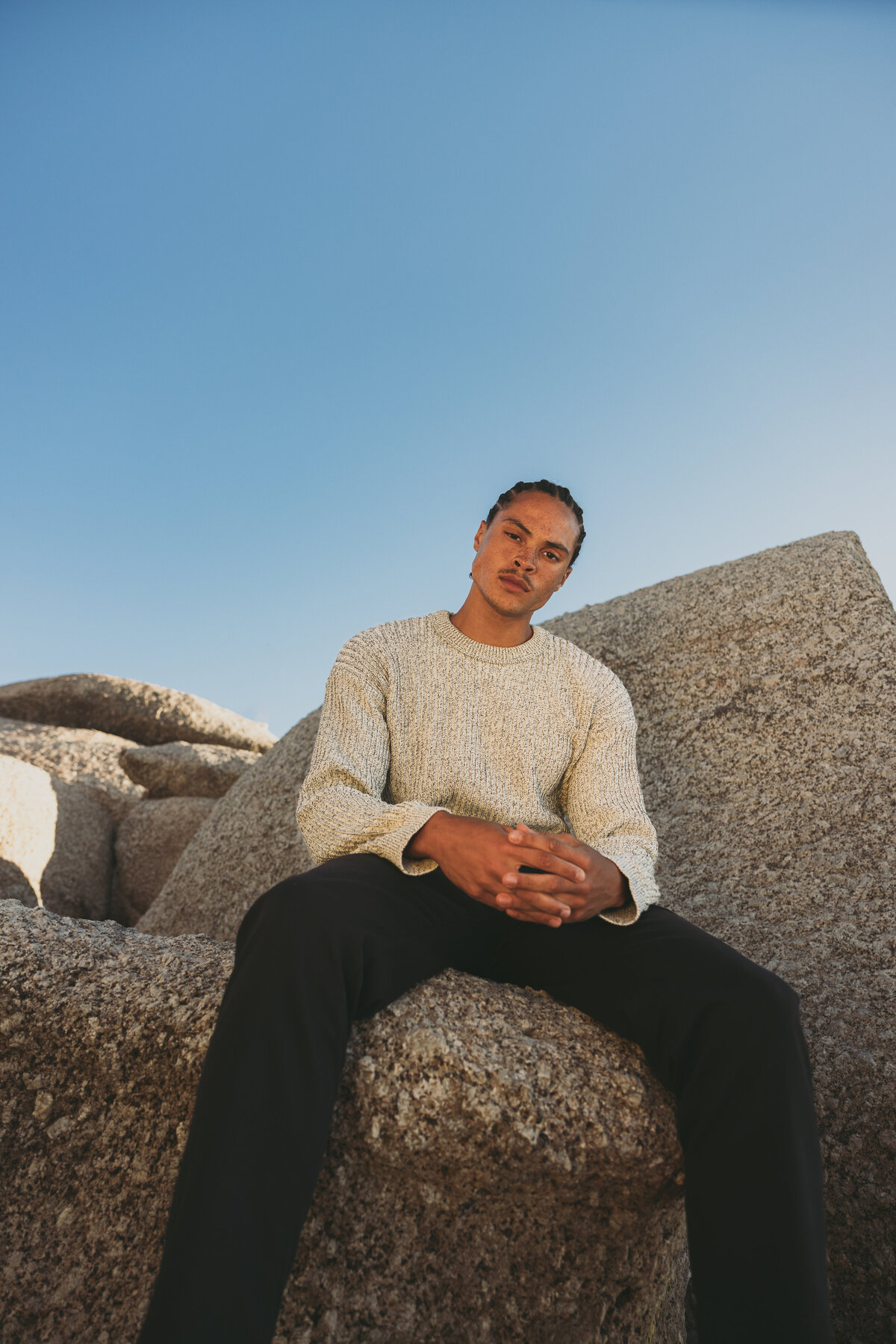 Portrait of Timothe sitting confidently on large rocks in Cape Town, wearing a light knitted sweater and black pants under a clear blue sky.