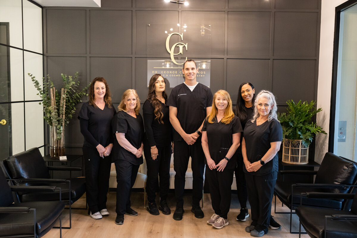 A team photo of Dr. George Christakos and the dental staff standing in front of their brand new office, featuring a black board and batten wall and modern, sophisticated interior design.