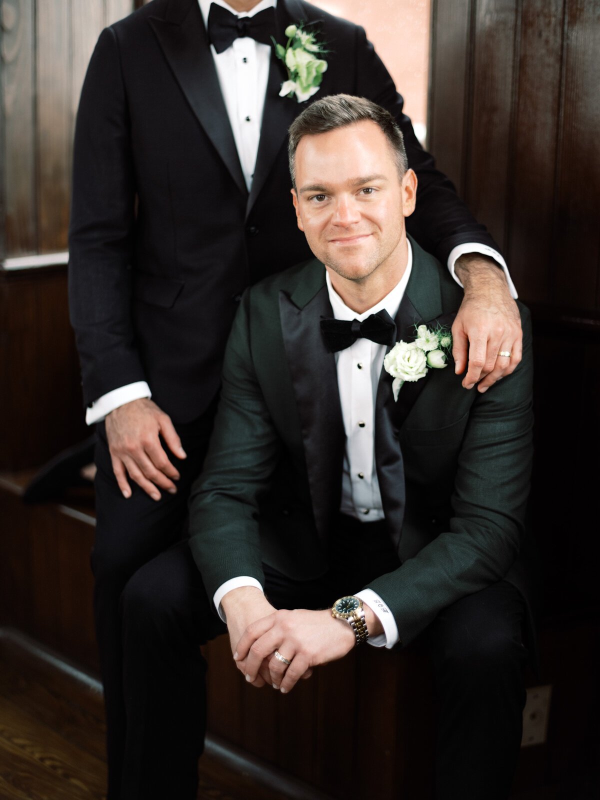 A groom seated elegantly with a charming smile, exuding confidence and joy, showcasing his striking outfit and the love that surrounds him on this wonderful day.