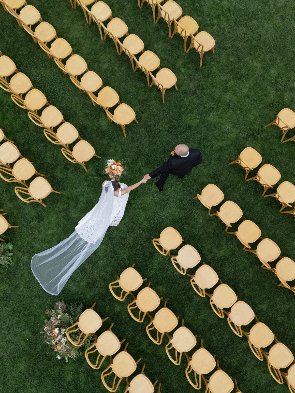 overhead drone shot of wedding couple walking down aisle of light wood chairs and florals representing full service design and calligraphy by Life in Letters.