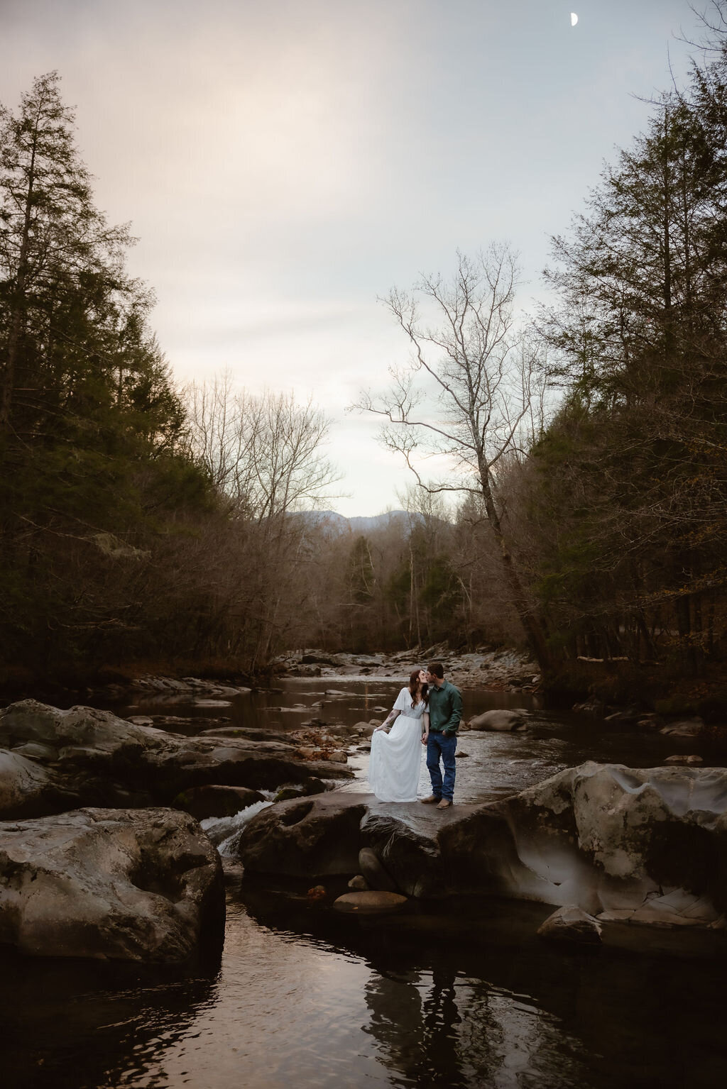 Bride and groom standing on a large rock beside the river at Greenbrier, sharing a kiss under a twilight sky with the moon visible, during their eloping to Gatlinburg ceremony in the Smoky Mountains.

