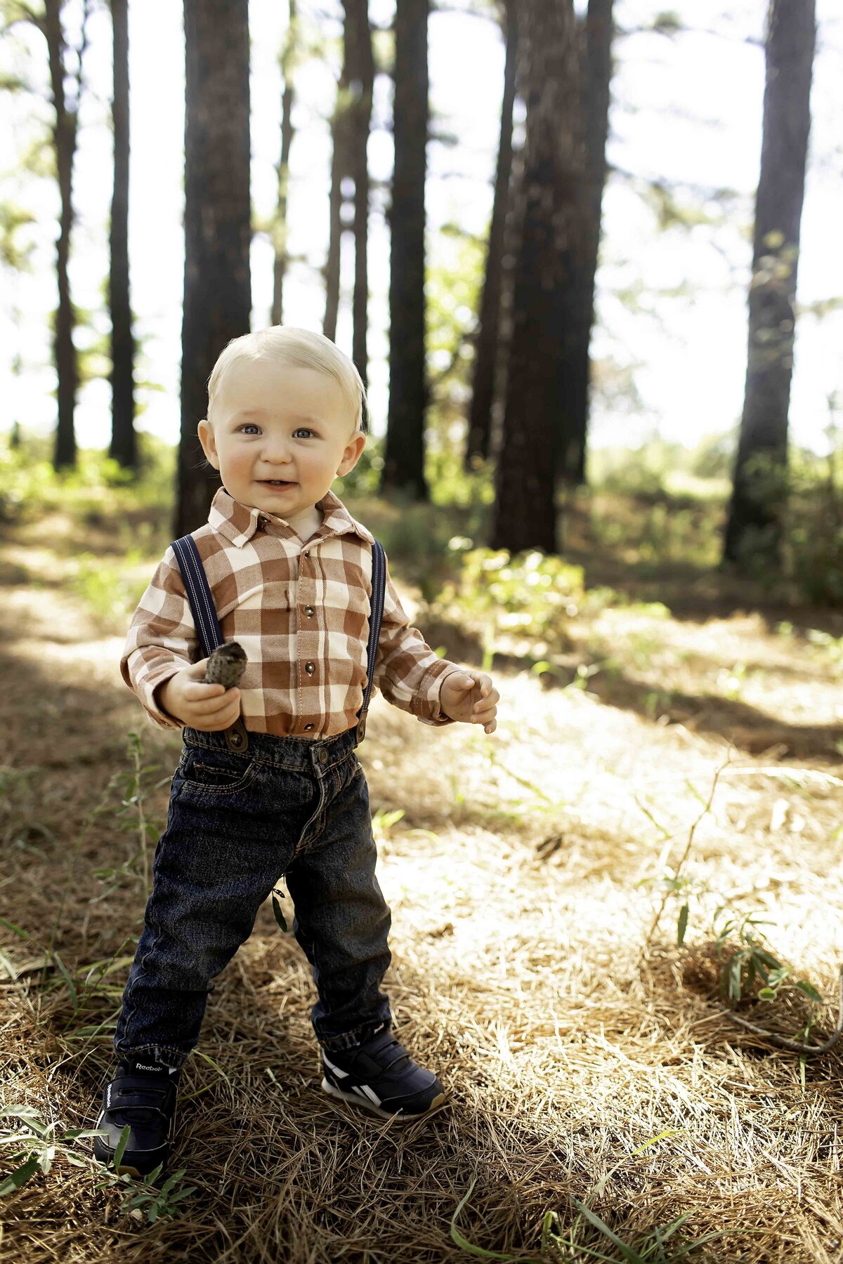 Birthday boy dressed in suspenders and overalls, standing in the pine trees, his hands on his hips