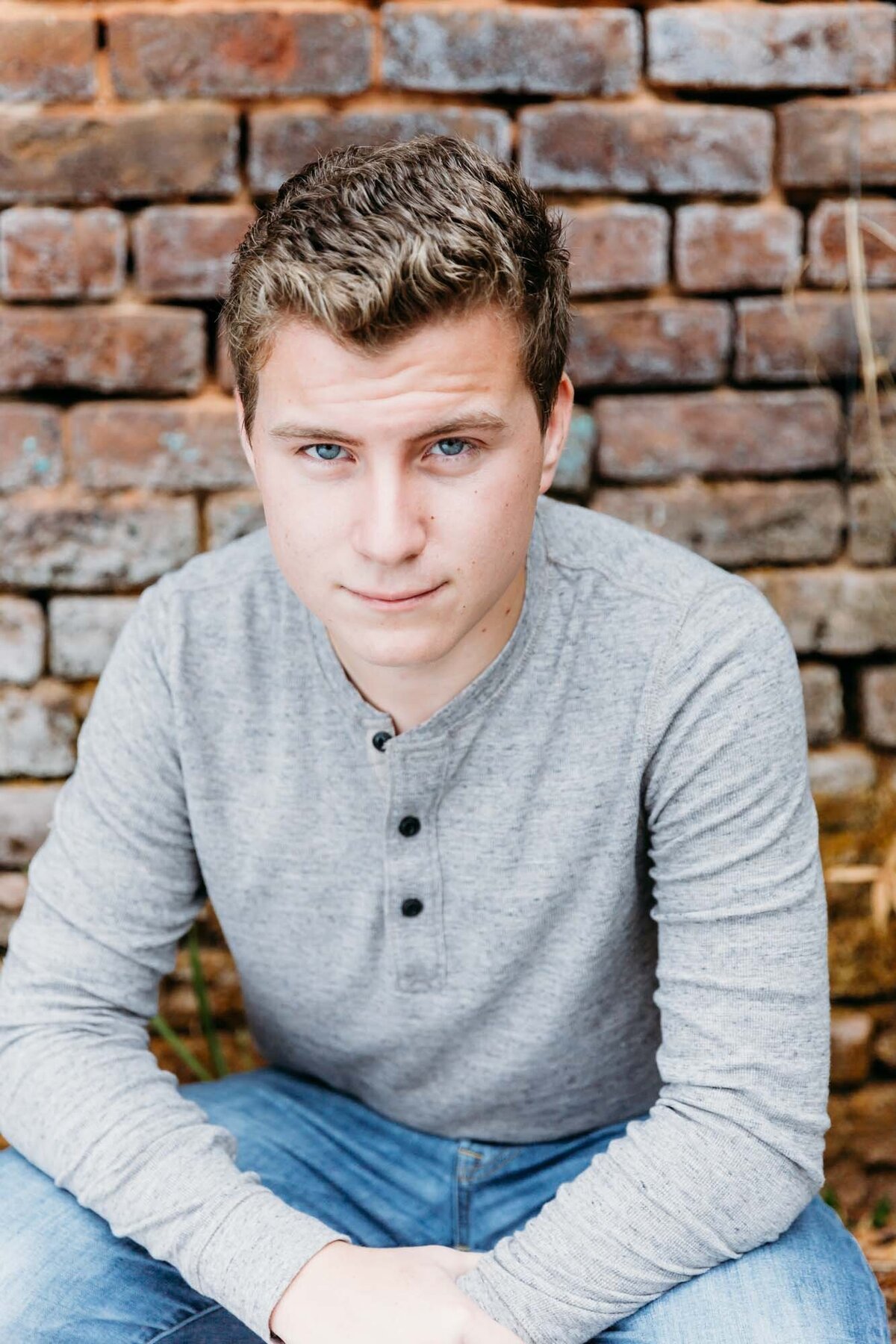 Senior portrait of a boy in a gray shirt in front of a brick wall.