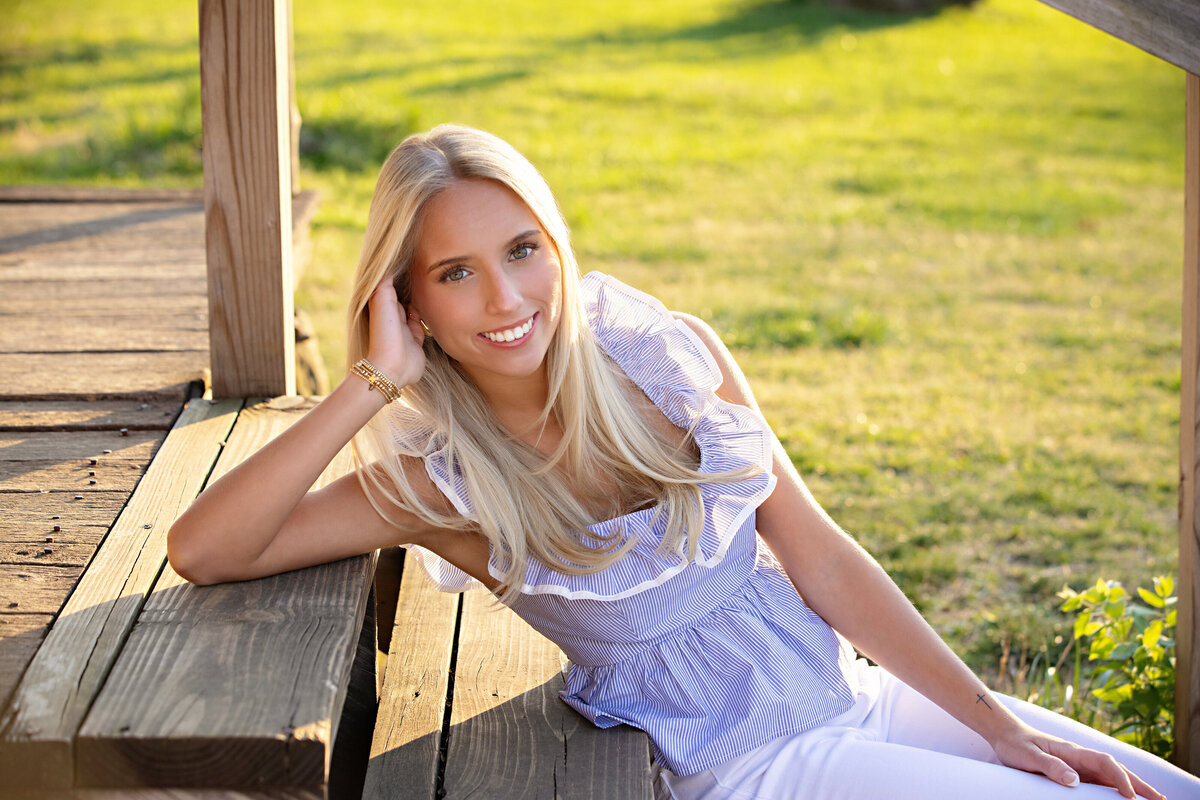 High school senior with blonde hair seated on wooden steps of a rustic cabin at Anne Springs Close Greenway in Fort Mill SC, wearing a ruffled top and relaxed expression in soft spring light