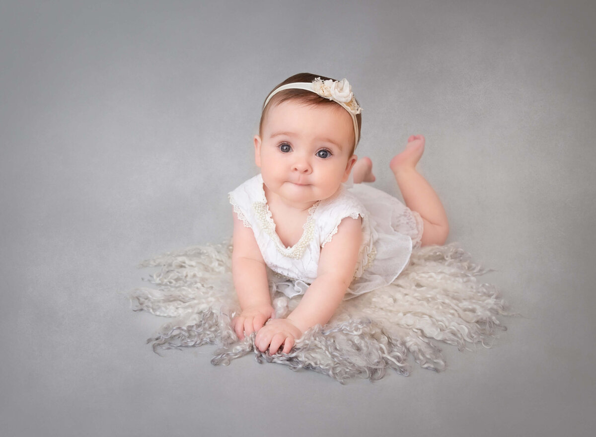 6-month-old baby girl lying on her belly with her arms folded, propped up on her elbows and smiling at the camera during a milestone photoshoot in NYC