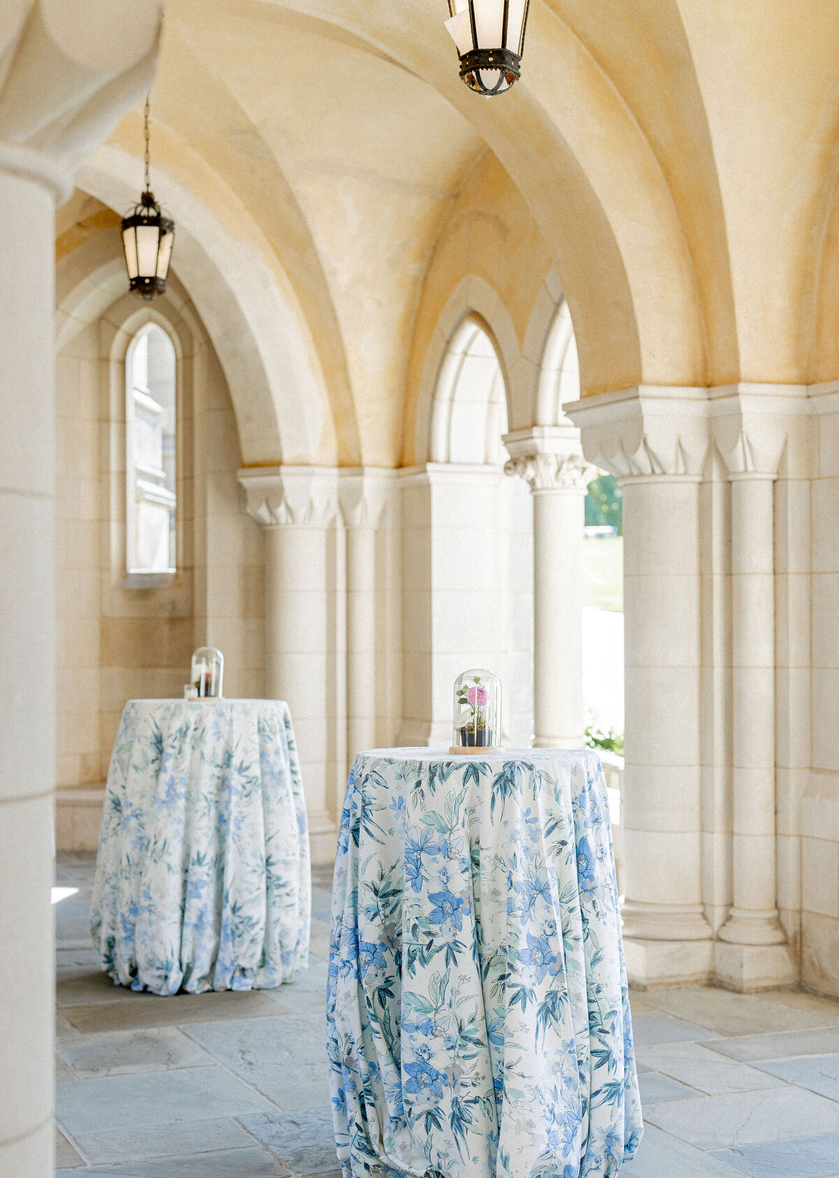 A stone arched hallway with two high tables covered in blue and white floral tablecloths. Each table has a glass dome with a small bouquet inside. The scene is well-lit by hanging lantern-style lights attached to the ceiling.