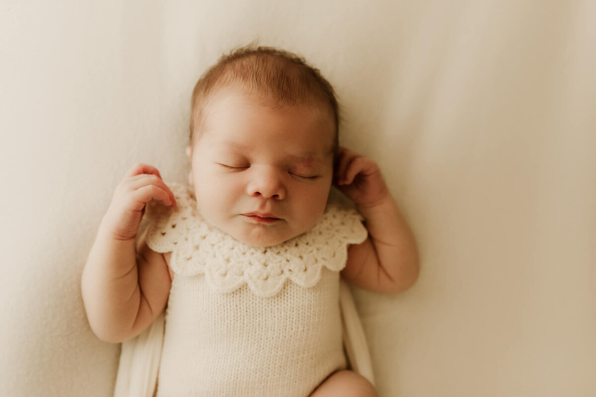Baby girl sleeps on a cream blanket while wearing a collared cream romper.