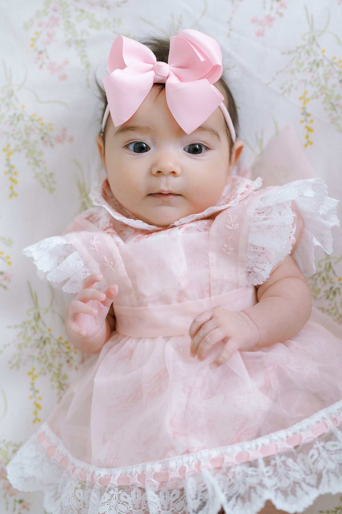 Newborn girl wtih eyes wide open wearing a lacy pink chiffon dress and laying on a floral blanket..