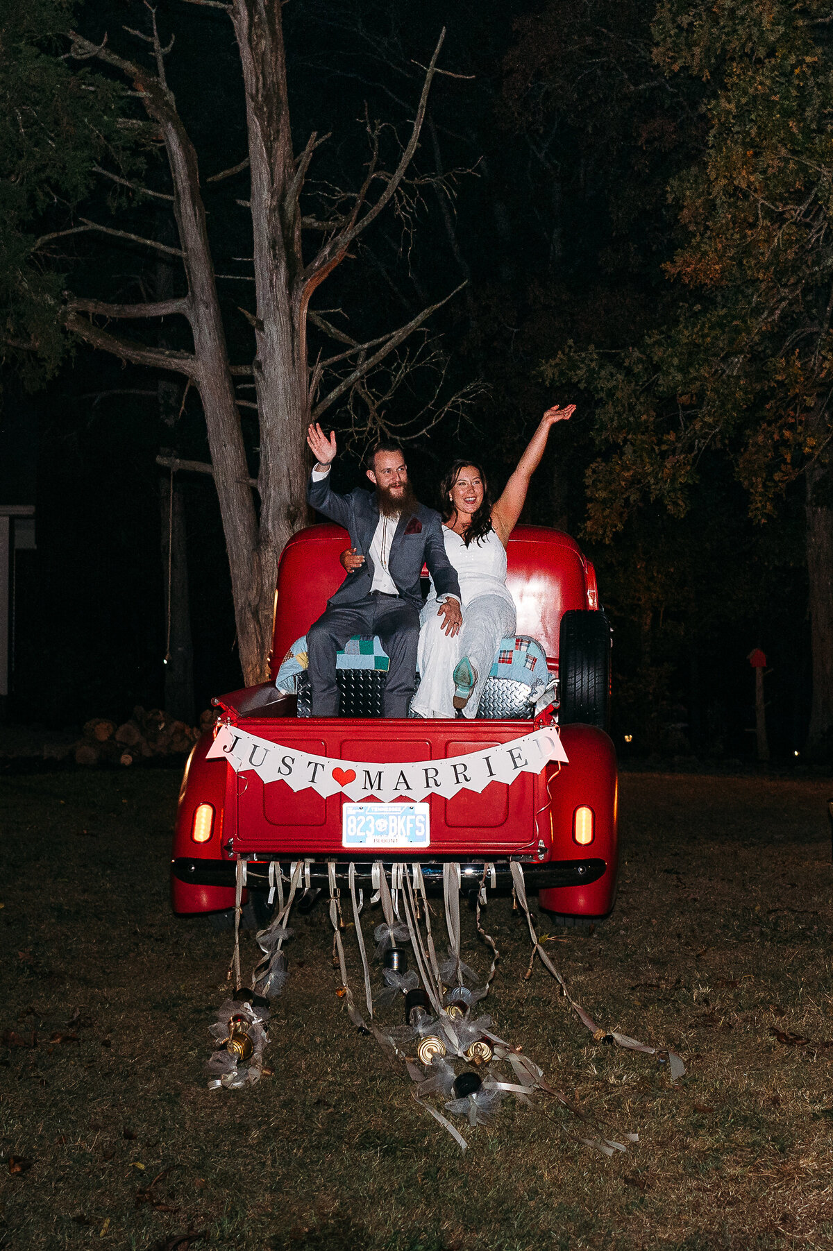 Bride and groom sitting on the back of a red “Just Married” truck at night with streamers hanging