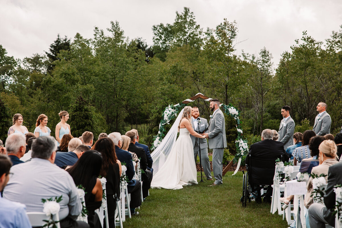 A peek down the aisle as a bride and groom hold hands under the arbor with guests watching